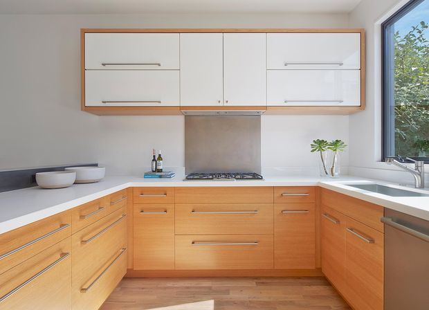 Bright, compact contemporary kitchen with an L-shaped timber-look cabinetry layout, white benchtops, white wall-mounted overhead cupboards, and long horizontal metal handles. A stainless-steel cooktop and rangehood sit beneath a pale splashback, with a sink positioned by the large corner window. Timber flooring and warm natural light create a clean, functional atmosphere.
