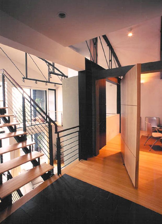 Interior view of a contemporary multi-level space with an open stairway featuring timber treads and black metal cable railings. Warm timber flooring continues through the hall, while a dramatic black structural or partition element frames the circulation area. Exposed dark metal framing, white walls, track lighting, and large glazed openings create an industrial loft-like character. A small seating or work area is visible to the right.