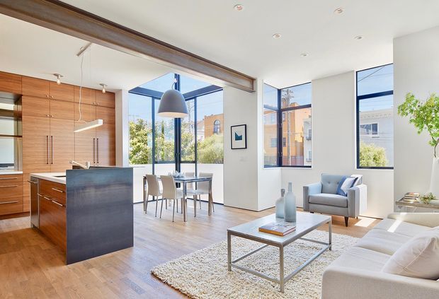 Bright open-plan contemporary interior combining a kitchen, dining area and living room. The space features warm timber cabinetry and flooring, a dark island, large black-framed windows, white walls, recessed lighting, a sculptural pendant, and minimalist furnishings. A substantial exposed timber beam separates the kitchen zone from the living area, with urban buildings visible outside.