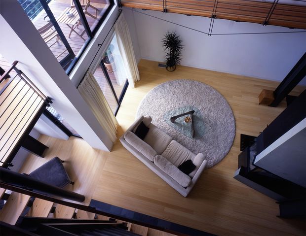 High-angle view into a bright double-height living room with pale timber flooring, a large round textured rug, curved light sofa, triangular glass coffee table, tall glazing with curtains, and dark steel-framed stair and mezzanine structures. A timber-lined mezzanine or upper-level walkway spans the top of the space, emphasizing the open multi-level layout.