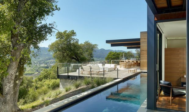 A contemporary hillside residence viewed from beside a narrow reflecting pool or swimming pool, with a large glass terrace, outdoor lounge and dining area, timber-lined soffits, and dark framed glazing. The home overlooks a verdant valley and mountain landscape in bright clear daylight.
