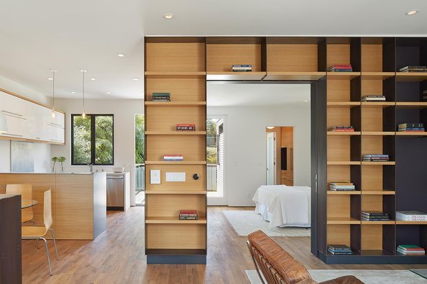 Open-plan residential interior with a timber kitchen and dining area on the left, and a full-height built-in timber and dark-framed shelving partition separating the living space from a bedroom visible through a large central opening. Warm timber flooring, recessed ceiling lights, pendant lights, bookshelves, and a white-covered bed create a clean, contemporary fit-out.