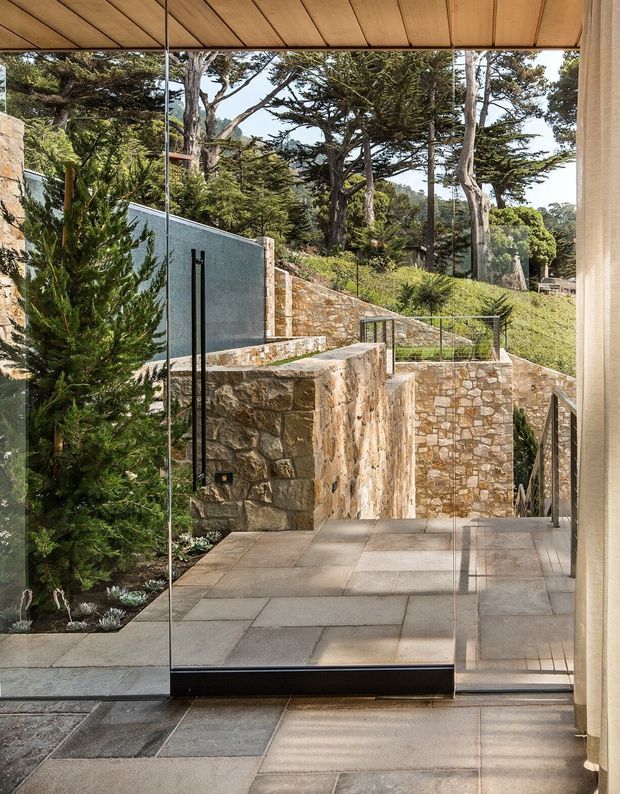 A view from inside through a large frameless glass door toward a landscaped, terraced outdoor area. The space features substantial rough-cut stone retaining walls and steps, large-format grey stone paving, dark metal balustrades, and mature trees beyond. A timber-lined ceiling and pale interior wall frame the glazed opening, creating a strong connection between the interior and the garden.