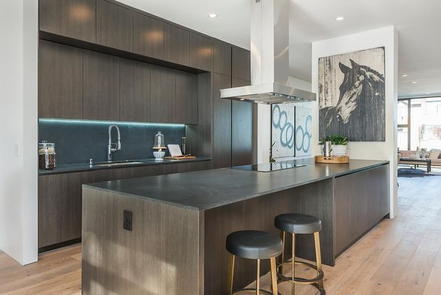 A contemporary open-plan kitchen dominated by dark timber cabinetry and a large central island. The kitchen features a dark stone-look benchtop, integrated appliances and storage, a stainless-steel undermount rangehood, a black splashback with under-cabinet lighting, and a brushed metal gooseneck tap. Two stools sit at the island, with a dining or living area visible beyond. Light timber flooring and white walls create contrast with the dark joinery.