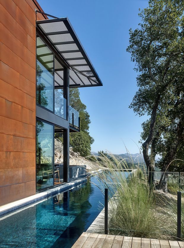 Exterior view of a contemporary multi-level residence beside a narrow swimming pool. The facade combines warm rust-coloured metal cladding with extensive floor-to-ceiling glazing and dark structural framing. A projecting translucent louvred or framed canopy shades the upper glazing, while a timber deck, glass balustrade, ornamental grasses, mature trees, rocky terrain and distant hills surround the pool.