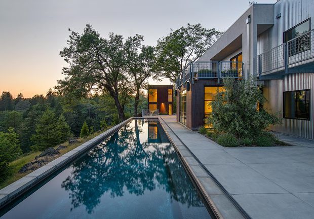 A contemporary multi-level house set beside a long narrow reflecting swimming pool, viewed at dusk. The building combines dark vertical metal cladding, concrete volumes, expansive glazing, balconies with metal railings, and warm interior lighting. Mature trees and dense landscape border the property, while the pool mirrors the surrounding trees and architecture.