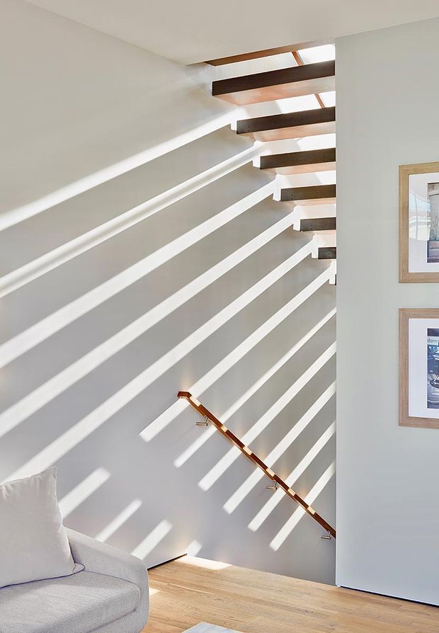Bright contemporary stairway with white walls and an open-riser staircase. Dark timber treads project from the wall, creating strong linear shadows and admitting abundant daylight from above. A slim timber handrail runs diagonally down the stair, with pale timber flooring and part of a light upholstered sofa visible at the lower left. Framed artwork is mounted on the adjacent wall.