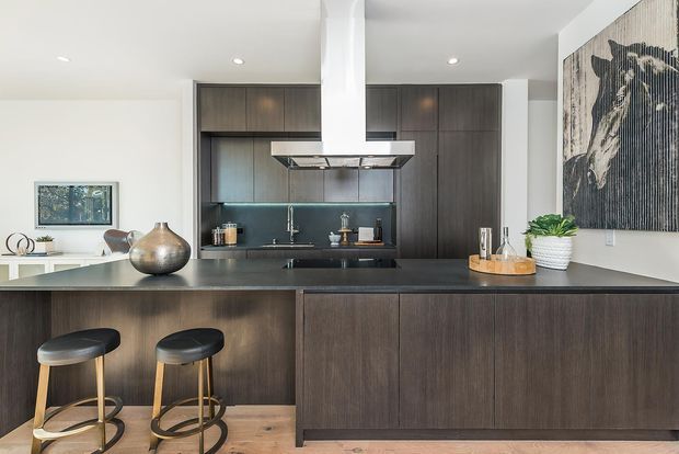 Contemporary open-plan kitchen dominated by a large dark wood island with two bar stools, dark full-height cabinetry, a black benchtop, and a matching dark splashback. A stainless-steel rangehood hangs above the cooking zone. The space has white walls and ceiling, timber flooring, integrated appliances, recessed ceiling lights, and a large monochrome artwork on the side wall. A living or dining area is partially visible beyond the kitchen.