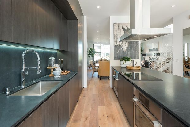 A contemporary galley-style kitchen with dark timber cabinetry, dark stone benchtops, an undermount sink, and a prominent stainless-steel spring faucet. A large central island with integrated cooktop and overhead stainless-steel rangehood faces an open dining and living area. Light timber flooring, white walls, recessed ceiling lights, and a glass balustrade create a bright, streamlined interior.