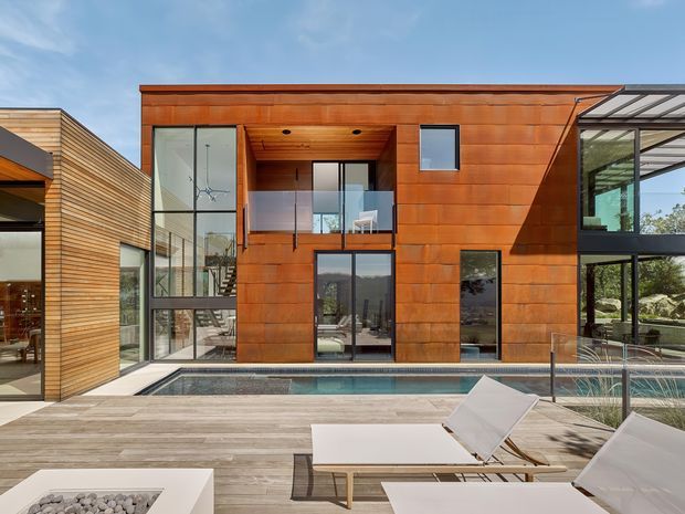 Contemporary multi-level residential exterior viewed from a pool terrace, featuring warm timber cladding, expansive dark-framed glazing, a recessed upper-level balcony, and a glazed pavilion-like wing. The foreground includes a long swimming pool, timber-look decking, glass balustrades, and outdoor lounge furniture with a square fire feature.