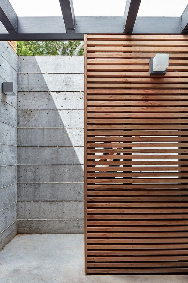 A modern semi-enclosed outdoor courtyard or walkway featuring a raw concrete block wall, a large horizontal timber slat privacy screen, exposed dark structural beams overhead, and concrete paving. The slatted screen partially reveals a timber stair or structure behind it. Wall-mounted rectangular light fittings and strong daylight create a minimalist architectural composition.