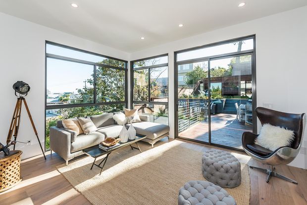 Bright contemporary living room with large corner windows and glazed doors opening to a timber deck. The room features a light sectional sofa, black leather lounge chair, tufted grey ottomans, a textured neutral rug, timber flooring, white walls and recessed ceiling lights. A leafy suburban outlook and neighbouring homes are visible through the glazing.