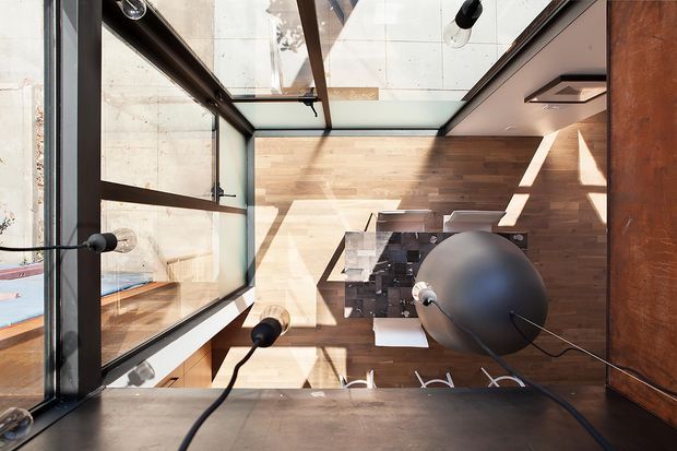 Overhead view of a contemporary kitchen and dining area, featuring warm timber flooring and wall cladding, dark-framed floor-to-ceiling glazing, a central dark stone island with stools, and distinctive black pendant lighting. Strong sunlight creates geometric shadows across the open-plan space.