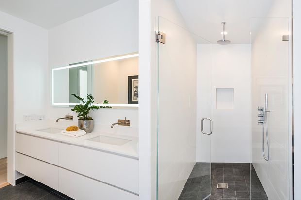 Bright contemporary bathroom featuring a wall-mounted double vanity with integrated white basins, brushed metal wall-mounted taps, a large illuminated mirror, and a separate frameless glass walk-in shower. The shower has white tiled walls, a handheld and overhead shower fitting, and dark grey tiled flooring. The palette is clean white with dark flooring and subtle metallic accents.