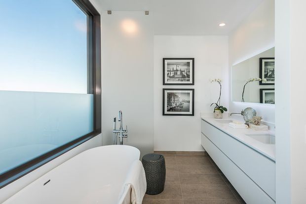A bright, contemporary bathroom featuring a freestanding white bathtub beside a large dark-framed window, a wall-mounted double vanity with a broad mirror, recessed ceiling lights, framed black-and-white artwork, and a small orchid. Large grey-brown floor tiles and predominantly white surfaces create a clean, refined atmosphere.