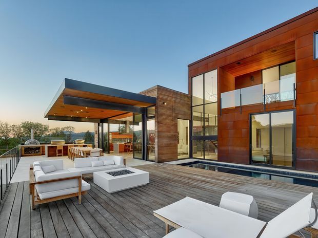 A large contemporary rooftop terrace and outdoor entertaining area adjoining a multi-level modern residence. The space features extensive timber decking, white outdoor lounge furniture, a dining table, glass balustrades, a covered outdoor kitchen and dining pavilion, floor-to-ceiling glazing, and warm timber and reddish-brown cladding.