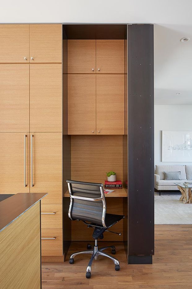 Built-in timber joinery with a compact integrated home office nook, featuring a desk, black leather office chair, overhead cupboards, tall storage cabinets, and dark contrasting side panels. The space opens toward a bright living room with a sofa and artwork, while timber flooring continues throughout.