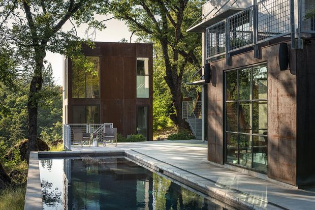 Contemporary multi-level residential property set among mature trees, featuring dark metal or timber-clad facades, extensive floor-to-ceiling glazing, projecting balconies, and a paved terrace beside a long rectangular swimming pool. The composition focuses on the building exterior, pool, and outdoor living terrace in a wooded setting.