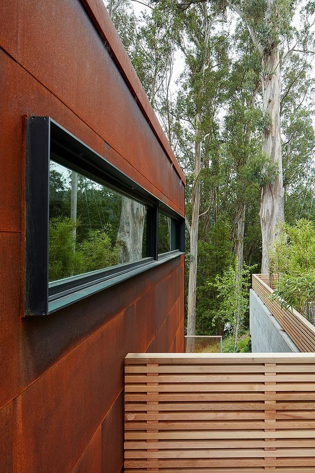 Close-up exterior architectural view of a contemporary building set among eucalyptus trees. The facade is clad in weathered rust-coloured steel panels with dark-framed horizontal glazing, beside a timber slatted privacy screen and elevated timber deck or walkway. Dense native vegetation surrounds the structure.