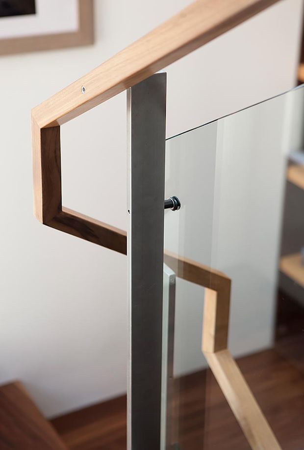 Close-up detail of a contemporary staircase balustrade featuring clear glass panels, slim dark metal supports and a warm timber handrail with angular joinery. The staircase is set against light painted walls with dark timber flooring.