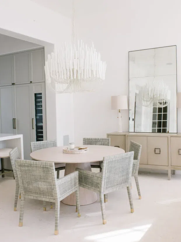 Bright, refined dining area with a round pale dining table surrounded by woven-back chairs. A large sculptural chandelier hangs above, while pale grey built-in cabinetry with integrated wine storage occupies the left side. A low sideboard with a large mirror and table lamp sits against the wall, creating an airy, layered interior with soft neutral finishes.