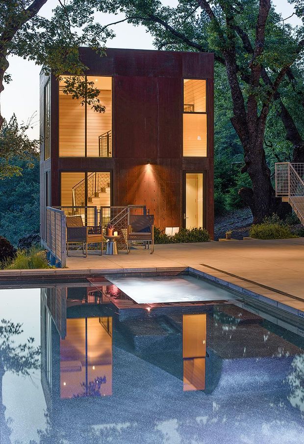 A compact, modern multi-storey pavilion set among mature trees beside a reflective swimming pool. The dark rust-toned metal-clad facade has large illuminated windows, visible stairways, a ground-level doorway, and a small outdoor terrace with seating. Pool reflections and warm interior lighting create a dramatic evening architectural photograph.
