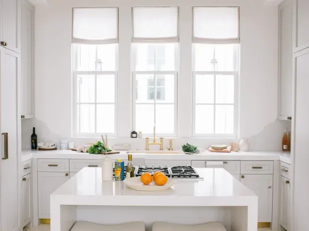 Bright, symmetrical white kitchen with three tall sash windows, shaker-style cabinetry, a central island with a white benchtop and integrated gas cooktop, and brass-toned tapware and hardware. The room has a clean, refined traditional feel with abundant natural light.