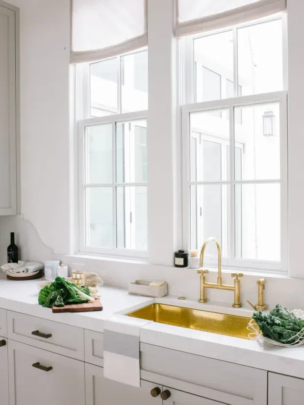 Bright, white traditional-style kitchen focused on a farmhouse sink beneath tall multi-pane windows. White Shaker cabinetry and white countertops create a clean backdrop, while polished brass/gold plumbing fixtures provide a strong accent. A dish towel, vegetables, and small countertop items are visible, with an airy glazed view beyond the windows.