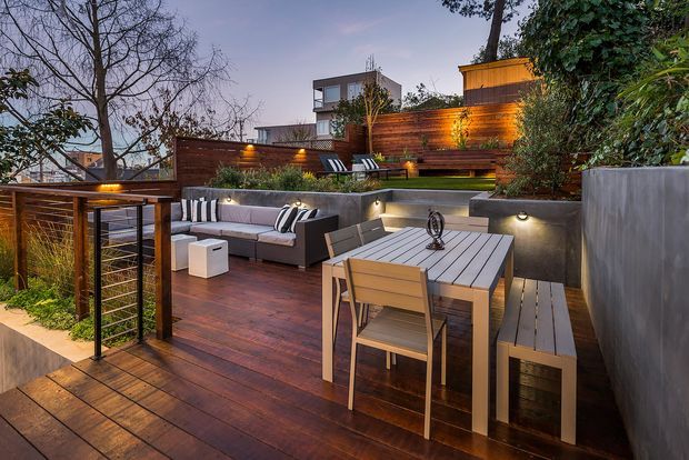 A landscaped multi-level timber deck and outdoor entertaining area at dusk. The space features a dining table with bench seating, an outdoor lounge with a sectional sofa and cushions, built-in concrete planters and retaining walls, warm integrated lighting, timber privacy screening, and metal-framed balustrading. Dense planting surrounds the elevated deck, with neighbouring homes and a modern multi-storey residence visible beyond.