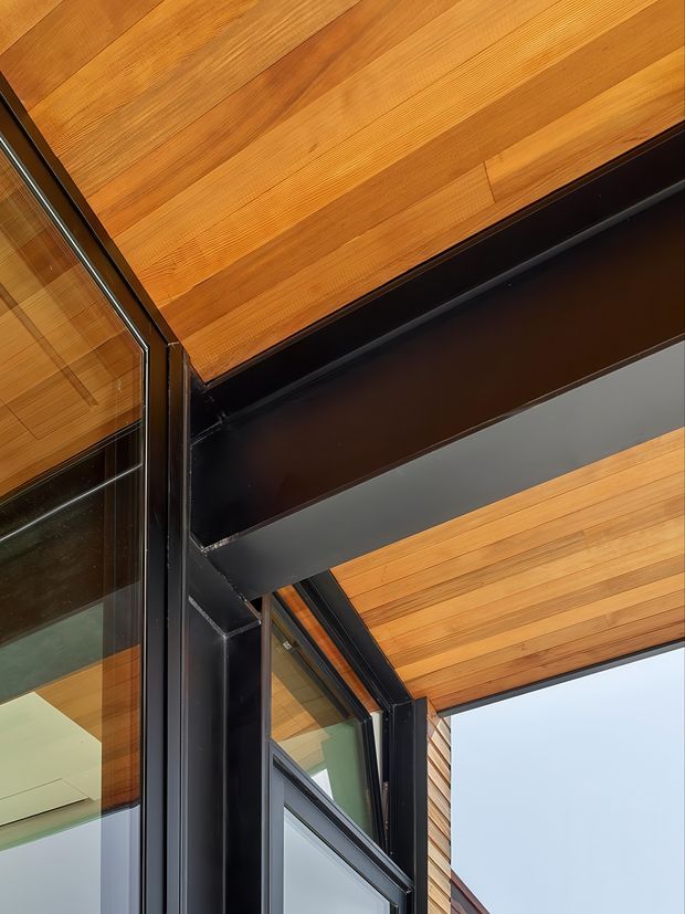 Close-up architectural detail of a contemporary building corner, showing warm timber soffit or ceiling boards, exposed dark metal structural beams, and large black-framed glazed openings. The image emphasizes the junction between timber cladding, steel framing, and glazing, with a small portion of pale sky visible beyond.