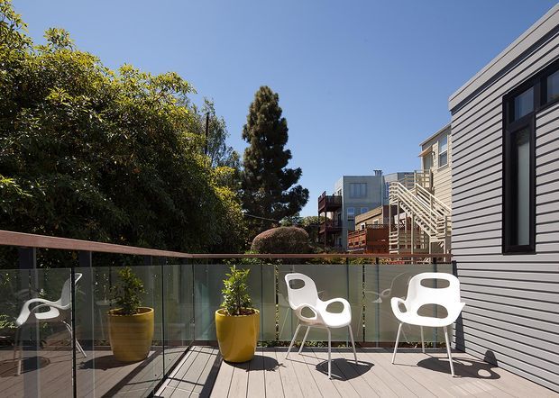 Sunny exterior view of a contemporary residential deck or balcony with pale timber-look decking, frameless glass balustrading, and white sculptural outdoor chairs. A grey horizontal-clad building facade with dark-framed windows occupies the right side, while mature greenery and neighbouring homes form the background. The space feels bright, open, and modern.