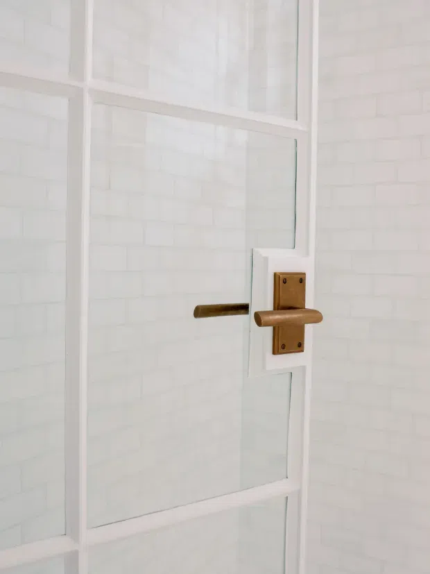 Close-up architectural detail of a white-framed glazed door or partition with a warm bronze/brass lever handle and rectangular backplate. White brick-pattern wall tiles are visible behind the glass, creating a clean, light bathroom-like setting, though the wider room is not shown.
