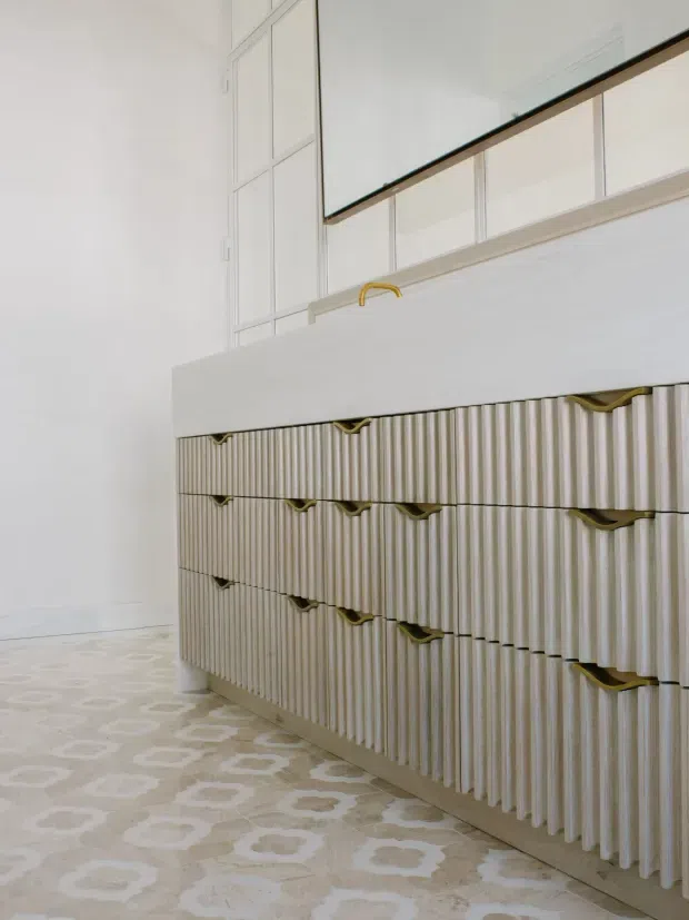 Bright close-up interior view of a refined built-in cabinetry unit, likely in a kitchen or utility space. The cabinetry has vertically fluted light-toned fronts with curved brass-gold pulls, a plain white benchtop, and upper white-framed glazed or mirrored cabinetry. A pale patterned floor adds a soft decorative element. The image emphasizes custom joinery and material detailing.