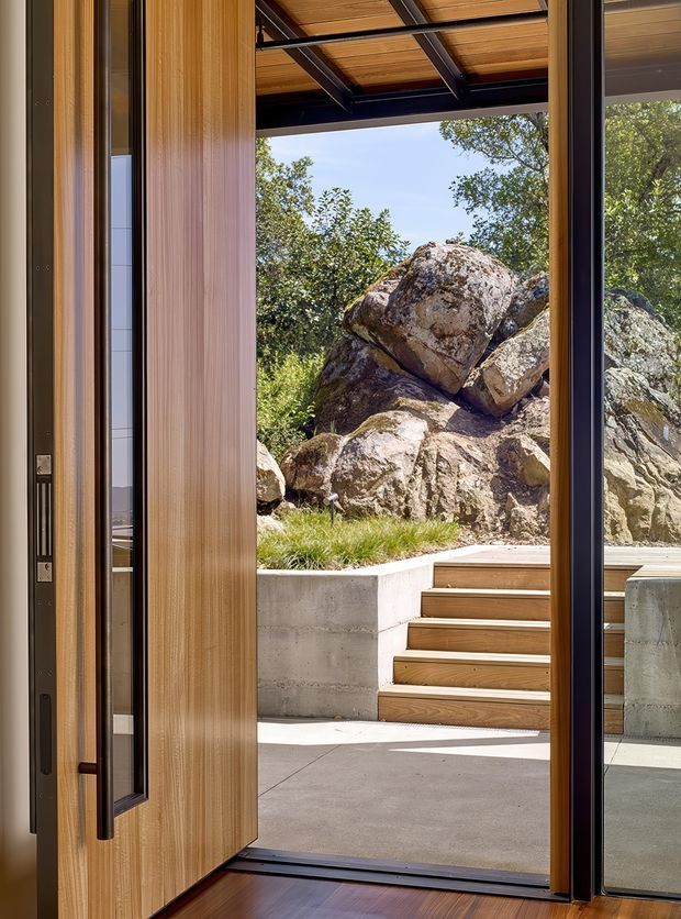 A contemporary indoor-outdoor threshold viewed from inside through a tall timber-framed glazed door. The opening leads to a concrete-paved courtyard or terrace with timber steps, concrete retaining walls, and a rugged natural rock outcrop surrounded by vegetation. A timber-lined overhead canopy with dark exposed steel framing extends over the exterior area. The image emphasises the strong connection between the interior and rocky landscape, with warm timber, black framing, concrete, and natural stone.
