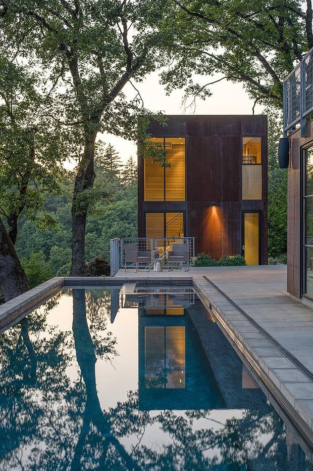 A modern multi-level house set among mature trees, viewed from beside a reflecting swimming pool. The dark timber-clad rectangular structure has large glazed openings glowing warmly from within, a small elevated terrace with metal railings, and a paved pool deck. Another glazed building volume is visible at the right edge, reinforcing the contemporary woodland setting.