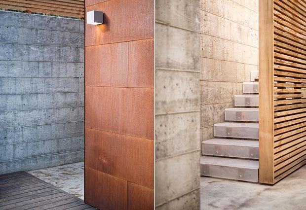 A contemporary architectural interior/exterior threshold featuring a tall rust-toned metal-clad door beside a textured exposed concrete wall. The view continues inside to a minimalist concrete staircase, with warm timber slatted screening and a small modern wall light providing contrast. Materials and detailing emphasize an industrial, refined aesthetic.