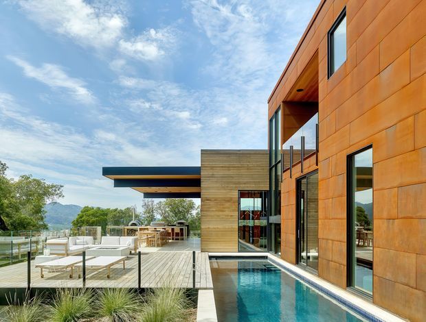 Exterior architectural photograph of a contemporary multi-level residence on a landscaped, elevated site. The building features extensive warm rust-toned metal cladding, horizontal timber wall sections, large dark-framed glazing, and strong projecting roof forms. A narrow swimming pool runs alongside the facade, with a timber deck, outdoor lounge furniture, glass balustrades, and distant mountain views.