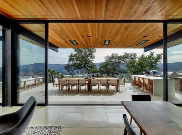 A contemporary indoor dining area opens through fully glazed black-framed sliding doors onto a covered outdoor dining terrace and outdoor kitchen. The space features a warm timber-lined ceiling with recessed lighting, large pale tiled flooring, dark window and door frames, and a long timber dining table with chairs. Mountainous landscape is visible beyond the terrace.