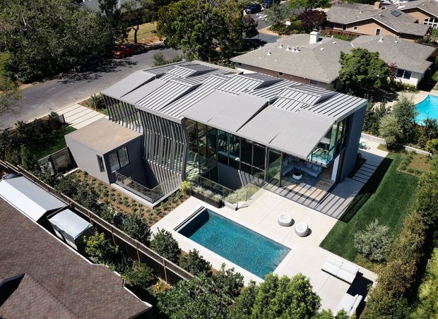 Aerial view of a contemporary multi-wing residential house with a distinctive ribbed metal roof and extensive floor-to-ceiling glazing. The home is arranged around a landscaped outdoor area with a rectangular swimming pool, paved terraces, lawns, garden planting, and outdoor seating. Neighbouring suburban homes and streets surround the property.