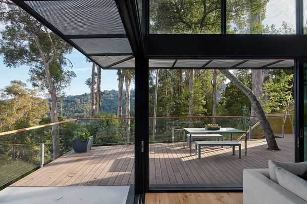 A contemporary indoor-outdoor living area opening onto a large elevated timber deck surrounded by mature trees. Floor-to-ceiling black-framed glazing and sliding doors connect the interior to the deck, which includes a glass balustrade, outdoor dining table and benches, and potted plants. The image has a natural bush setting, warm timber surfaces, and strong visual connection to the landscape.