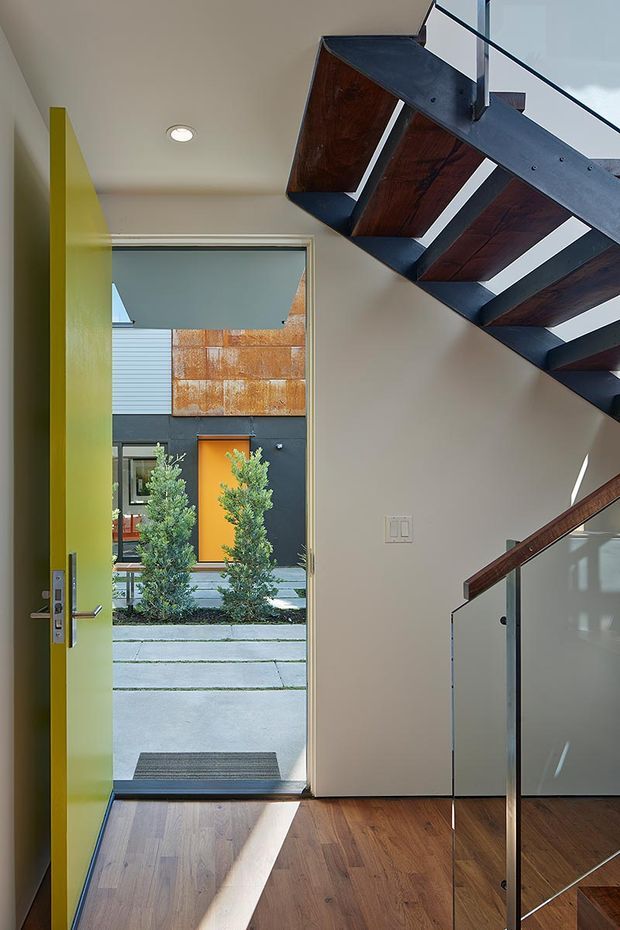 A modern residential entryway viewed from inside, with an open lime-green front door, warm timber flooring, and a dramatic exposed staircase overhead. The staircase combines dark timber treads with dark metal stringers and a clear glass balustrade. Through the doorway, a contemporary courtyard and neighbouring facade with rust-toned cladding are visible. Strong daylight creates a bright, welcoming interior with contrasting natural and industrial materials.