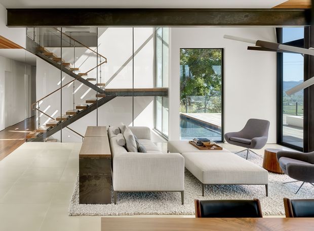 Bright, double-height contemporary living room with a sculptural open-riser staircase, glass balustrades, exposed dark structural beams, and extensive floor-to-ceiling glazing. A large pale sectional sofa and ottoman sit on a textured rug, with mid-century-inspired lounge chairs, a low coffee table, and a dining table in the foreground. The room opens visually to a pool and landscape beyond the windows.