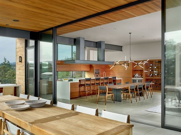 A spacious open-plan kitchen, dining, and bar area viewed through extensive floor-to-ceiling glazing. The room features extensive warm timber lining on the ceiling and walls, a large kitchen island with bar seating, dark structural steel elements, concrete-toned flooring, integrated appliances, and a wine display wall. A substantial timber dining table occupies the foreground, while sculptural contemporary pendant lighting hangs over the dining area. The glazing opens toward a leafy landscape and creates a strong indoor-outdoor connection.