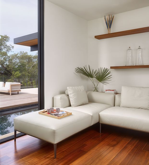Bright, refined living room with a cream sectional sofa and ottoman arranged beside full-height glazing. The room features warm polished timber flooring, white walls, slim dark window framing, and simple floating timber shelves displaying glass vessels and decorative branches. Through the glazing, a timber deck, outdoor seating, and a reflecting pool are visible.