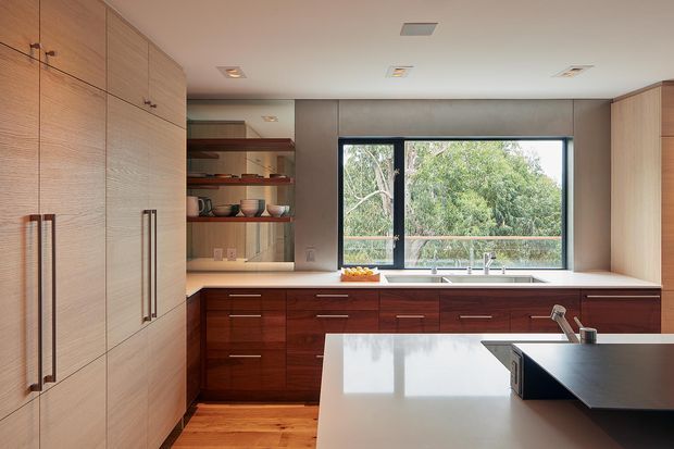 Contemporary kitchen with extensive timber cabinetry, a dark timber island base, pale stone benchtops, integrated shelving, and a large black-framed window overlooking greenery. The room has recessed ceiling lighting and a warm, refined material palette.