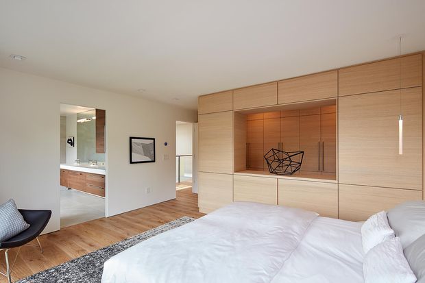 A warm, contemporary bedroom with a large built-in light timber wall unit spanning the far wall. The joinery includes flush cabinetry, an illuminated recessed display niche, and integrated vertical handles. A bed with white bedding occupies the foreground, accompanied by a dark patterned rug and a black lounge chair. An open doorway reveals a bathroom or dressing area with matching timber cabinetry, while a minimalist pendant light hangs beside the joinery.