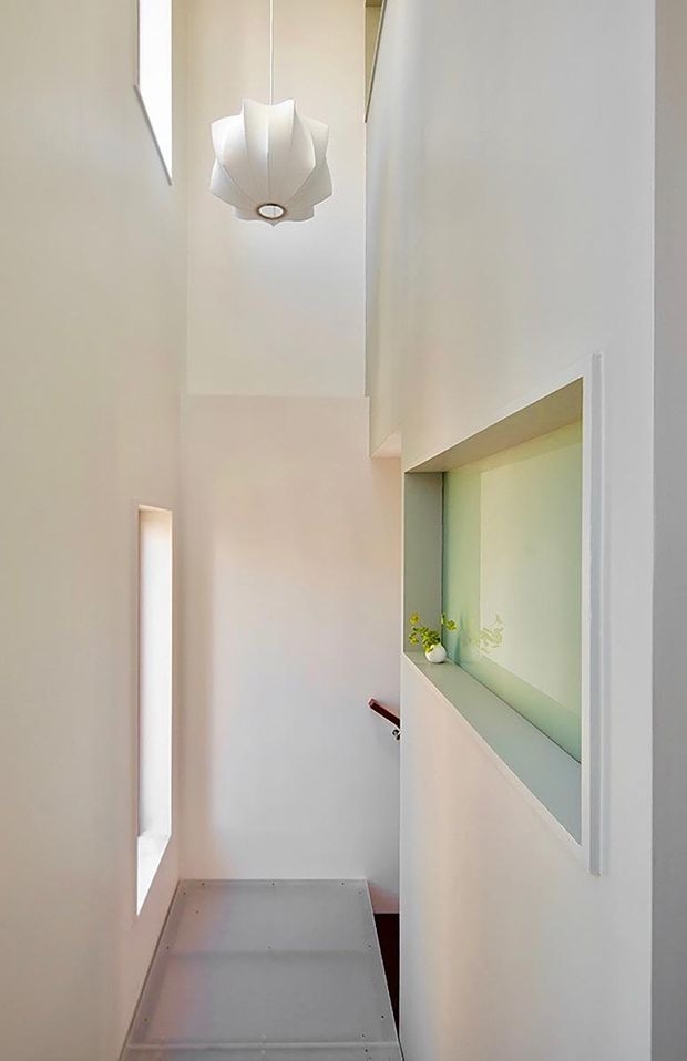 A narrow, minimalist interior passage or stair landing with very high ceilings, white walls, and strong vertical geometry. A sculptural white pendant light hangs centrally, while tall narrow glazing or openings bring in daylight. A recessed wall niche with a pale greenish finish contains a small potted plant, and a partial stair or landing edge is visible below.