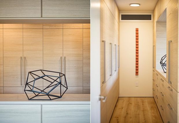 A warm, refined interior showing built-in light timber cabinetry and drawers surrounding a narrow hallway or dressing corridor. The left side features a geometric black wire sculpture on a counter, while the right side includes a large mirror reflecting the sculpture and cabinetry. A vertical orange decorative wall feature and recessed ceiling light add contrast. The space has a timber floor, light neutral walls, and integrated ventilation above the doorway.