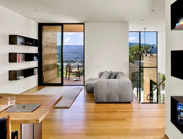 Bright contemporary living room with warm timber flooring and a substantial timber coffee table, a large grey sectional sofa, and built-in open shelving with dark accents. Full-height glazing and a sliding glass door open to a balcony or terrace with expansive mountain views. A stairwell and wall-mounted television are visible on the right, while recessed ceiling lights keep the space clean and modern.