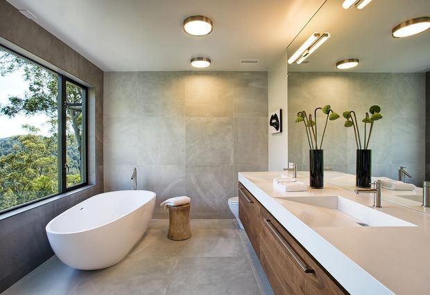 Contemporary bathroom with a freestanding white oval bathtub beside large dark-framed windows, a double vanity with timber cabinetry and white benchtop, full-height grey concrete-look wall and floor finishes, a large mirror, and integrated modern lighting. The windows overlook trees and a hilly landscape.